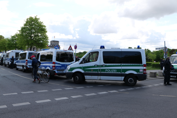 Eine Gruppe von Polizeiwagen entlang einer Straße geparkt mit einem Radfahrer im Vordergrund und Gebäuden, Bäumen und Schildern im Hintergrund unter einem bewölkten Himmel.
