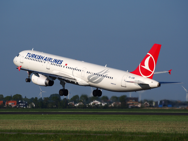 Ein Airbus A330-300 der Turkish Airlines startet vom Frankfurter Flughafen, mit Gras darunter, Gebäuden und Bäumen im Hintergrund und Windrädern in der Ferne unter einem klaren blauen Himmel.