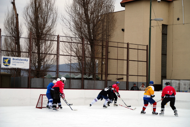 Menschen beim Eishockey auf einem Eisstadion mit Gebäuden, Bäumen, einer Straßenlaterne, einem Namensschild und Zäunen im Hintergrund unter einem Himmel.