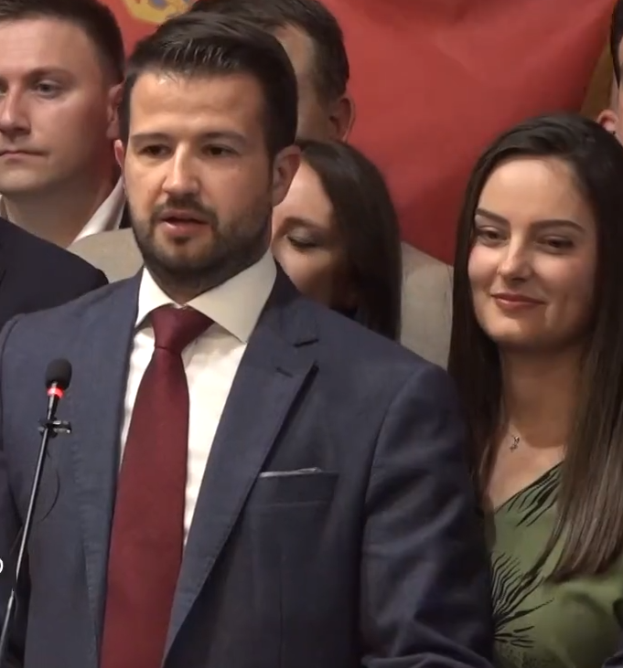 Ein Mann im Anzug und Krawatte steht an einem Rednerpult mit einem Mikrofon, spricht zu einer Menge mit einem Banner im Hintergrund, wahrscheinlich in Reaktion auf ein Regierungshandeln.