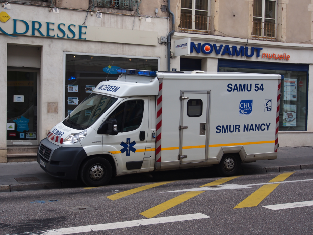 Ein Rettungswagen steht auf der Straße vor einem Krankenhausgebäude mit Fenstern, Geländern, Texttafeln, Plakaten und anderen Gegenständen.