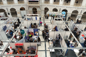 Eine große Gruppe von Menschen versammelt sich in einem Raum mit Ständen, Bannern, Tischen, Stühlen und Ballons, mit einem Gebäude im Hintergrund, das Bögen, Säulen, Geländer und einen Bildschirm aufweist.