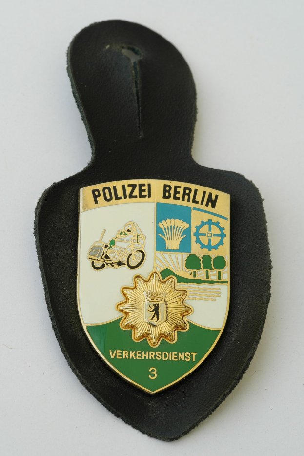 Ein schildförmiges Polizeibadge mit "Polizei Berlin" in schwarzer fetter Schrift, das ein weißes Stern auf blauem Hintergrund und einen weißen Rand hat, auf einer Oberfläche platziert.