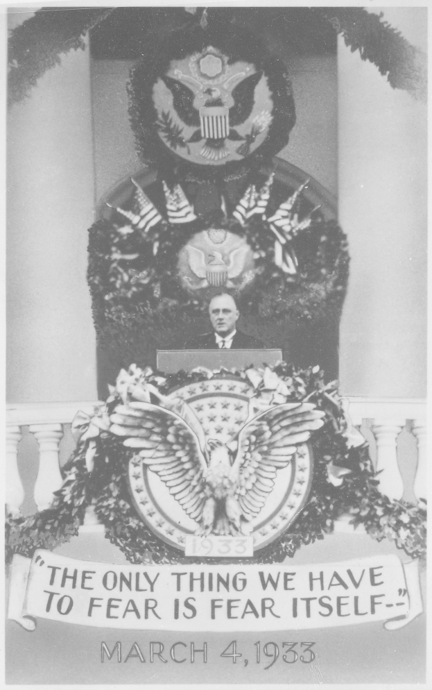 Schwarzes und weißes Foto eines Mannes, der an einem Rednerpult spricht, mit einem Kranz und einem Emblem an der Wand dahinter und dem Zitat "Das einzige, wovor wir uns fürchten müssen, ist die Furcht selbst" unten.