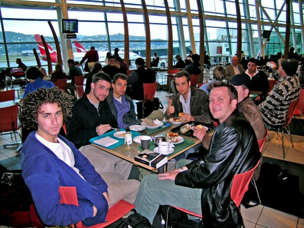 Eine Gruppe von Menschen sitzt um einen Tisch mit roten Stühlen in einem Flughafen, isst und unterhält sich, mit einer Glaswand, einem Hügel, einem Flugzeug und einem Fernseher im Hintergrund.