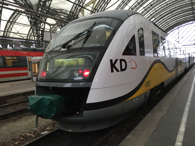 Ein weißer und gelber Zug parkt an einem Bahnhof mit einer Plattform auf der rechten Seite, ein weiterer Zug im Hintergrund und Metallstangen und ein Schuppen oben.