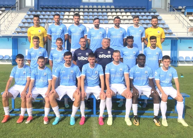 Eine Gruppe von Männern, einige sitzen auf Bänken und andere stehen auf einem Fußballfeld, mit Tribünen und Strukturen im Hintergrund.