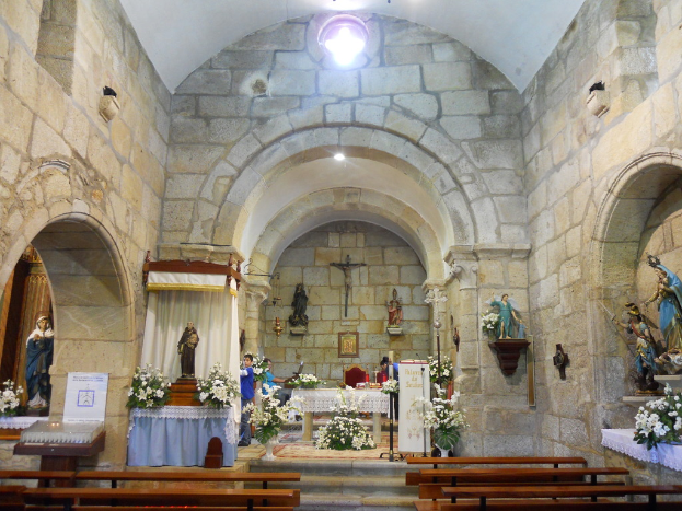 Innenansicht einer Kirche mit verschiedenen Skulpturen und Topfpflanzen auf leeren Holzbänken.