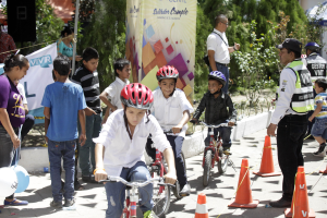 Eine Gruppe von Kindern, die auf Fahrrädern eine Straße entlangfahren, mit Verkehrskegeln, einige tragen Helme, andere stehen in der Nähe, mit einem Banner, Bäumen und Gebäuden im Hintergrund.