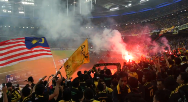 Eine Zuschauermenge in einem Stadion mit Fackeln, Fahnen und einer Kamera, mit Rauch und Schildern, die "Malaysia vs Malaysia Live Streaming" lesen.