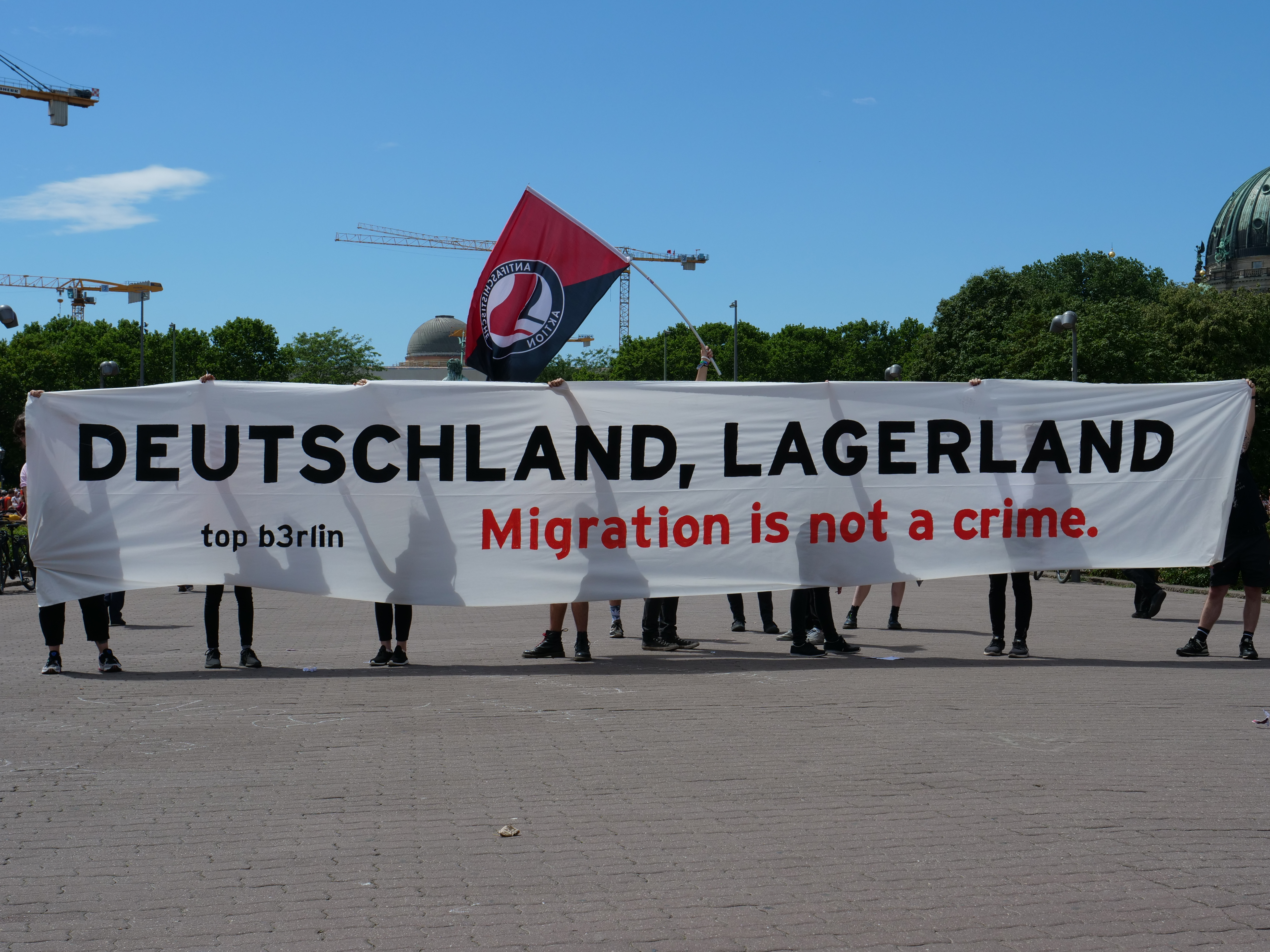 Eine Gruppe von Menschen hält ein Transparent mit der Aufschrift 'Deutschland, Lagerland Migration ist kein Verbrechen' und eine Flagge und steht auf dem Boden mit Straßenlaternen, Bäumen, Kränen, einem Gebäude und einem bewölkten Himmel im Hintergrund.
