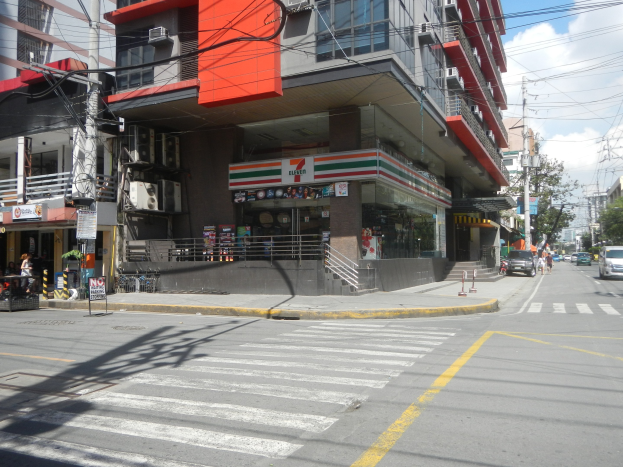 Eine belebte Stadtstraße mit Fahrzeugen, Fußgängern, hohen Gebäuden, einem 7-Eleven-Laden, Bäumen, Strommasten und einem bewölkten Himmel.