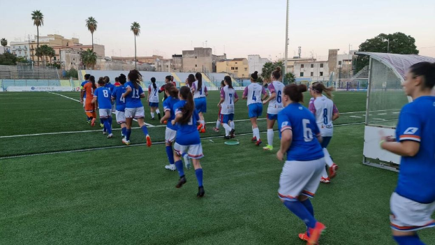 Eine Gruppe von Frauen, die Fußball auf einem Feld spielen, umgeben von Gebäuden, Bäumen und Pfählen unter einem klaren blauen Himmel, mit einem Torpfosten und Netz auf der rechten Seite.