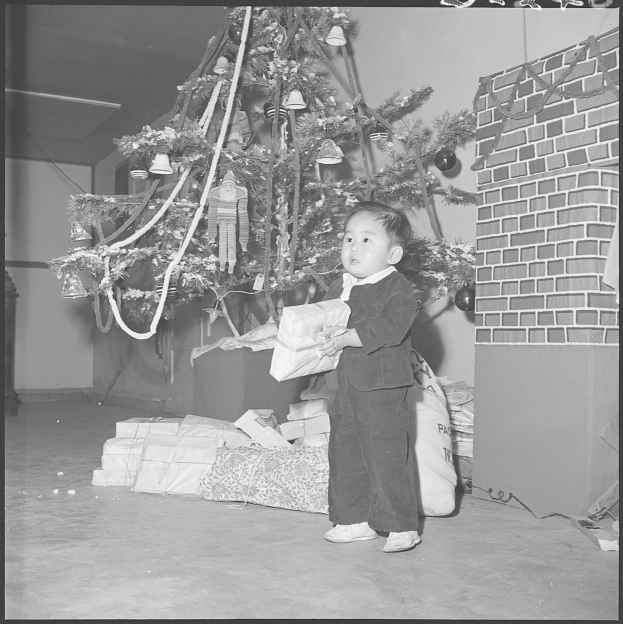 Ein junger Junge steht vor einem geschmückten Weihnachtsbaum, hält ein Geschenk in der Hand, mit mehreren anderen Geschenken um den Baum herum.