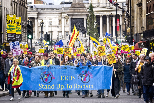 Eine Gruppe von Menschen marschiert mit einer 'Steh Auf gegen Rassismus'-Schlagzeile die Straße entlang, mit Gebäuden, Bäumen, Laternenpfählen, Verkehrsampeln und einer Statue in der Hintergrund unter einem bewölkten Himmel.