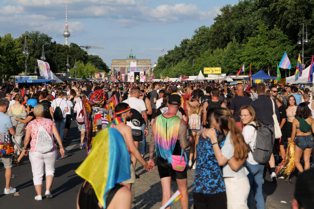 Eine große Menschenmenge geht eine Straße in Berlin, Deutschland, entlang, viele tragen Taschen und halten Fahnen, mit Zelten an den Seiten, Bäumen, Laternenmasten, einem Bogen und einem Wolkenhimmel im Hintergrund.