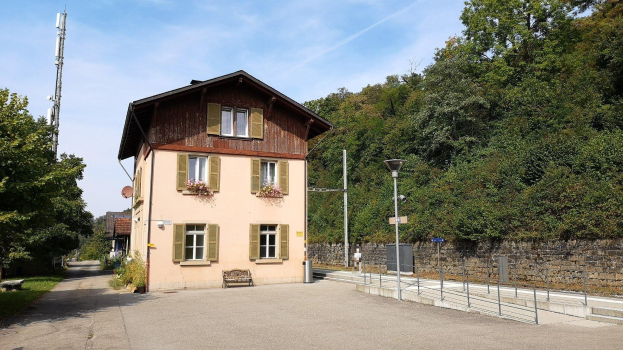 Ferienhaus zum Verkauf in Baden-Württemberg, Deutschland, mit einem mehrfenstrigen Gebäude mit einer vorderen Einfriedung, umgeben von Straßenmöbeln, Grünflächen und einem bewölkten Himmel.