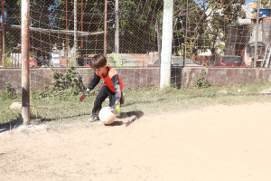 Junge auf einem Schmutzplatz Fußball spielend mit umgebenem Gras, Pflanzen, Pfählen, einem Zaun, einer Wand, Bäumen, Fahrzeugen, Gebäuden und einem bewölkten Himmel, trägt Schuhe.