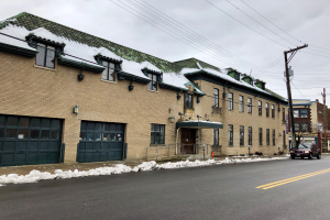 Großes Ziegelgebäude mit grüner Dachfläche an einer Straßenkreuzung, umgeben von Fahrzeugen, Schnee, Strommasten, Bäumen und einem bewölkten Himmel, identifiziert als die ehemalige Feuerwache St. Louis.