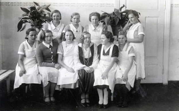 Eine Gruppe von Frauen sitzt auf einer Bank in einem Raum mit einer Wand, einer Tür und Zimmerpflanzen im Hintergrund.