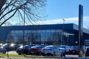Ein Mercedes-Autohaus mit parkenden Autos davor, umgeben von einem Metallzaun, Gras, einem Schild, Straßenlaternen, einer Baumgruppe und einem bewölkten Himmel.