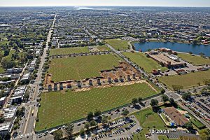 Luftaufnahme einer Stadt mit Gebäuden, Bäumen, Straßen, Fahrzeugen, Wasser und Wolken, die einen zentralen Platz als Parkstelle zeigt.