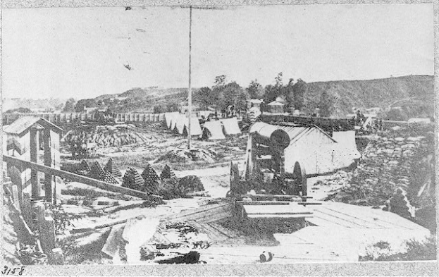 Schwarzes und weißes Foto einer Baustelle mit einer Maschine im Vordergrund, Schuppen, Bäumen, Hügeln und Himmel im Hintergrund, mit Text unten, der "Die Konstruktion einer neuen Brücke in der Stadt San Francisco, Kalifornien." lautet.