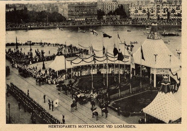 Ein altes Schwarz-Weiß-Foto einer Menge, die sich um einen Karneval mit Zelten, Fahnen, Stangen und Pferden versammelt hat, und im Hintergrund Gebäude, Bäume und Wasser zu sehen sind, mit Text am unteren Rand des Bildes.