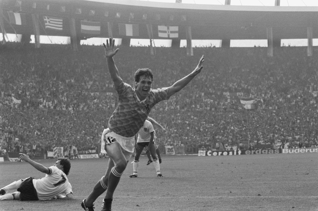 Schwarzes und weißes Foto eines Fußballspielers, der ein Tor feiert, umgeben von Teamkollegen und Stabmitarbeitern, mit Stadiontafeln, Fahnen und Pfosten im Hintergrund.