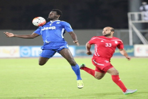 Zwei Männer beim Fußballspielen auf einem Feld, einer trägt ein blaues T-Shirt und Shorts, der andere ein rotes Kleid, im Hintergrund sind Schilder mit Text, ein Geländer, ein Pfosten und stehende Menschen zu sehen.