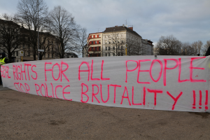 Eine Gruppe von Menschen, die auf dem Boden stehen und eine Fahne halten, auf der steht "Rechte für alle Menschen Stoppt Polizeigewalt", mit einem Straßenpfahl, einem Schild, Bäumen, Gebäuden mit Fenstern und einem bewölkten Himmel im Hintergrund.