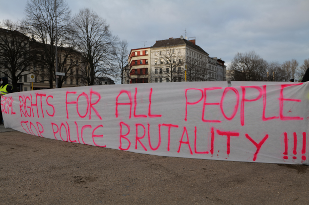 Eine Gruppe von Menschen, die auf dem Boden stehen und eine Fahne halten, auf der steht "Rechte für alle Menschen Stoppt Polizeigewalt", mit einem Straßenpfahl, einem Schild, Bäumen, Gebäuden mit Fenstern und einem bewölkten Himmel im Hintergrund.
