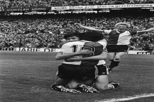 Drei Fußballspieler umarmen sich auf dem Boden, feiern ein Tor vor einer jubelnden Menge in einem schwarz-weißen Stadion.