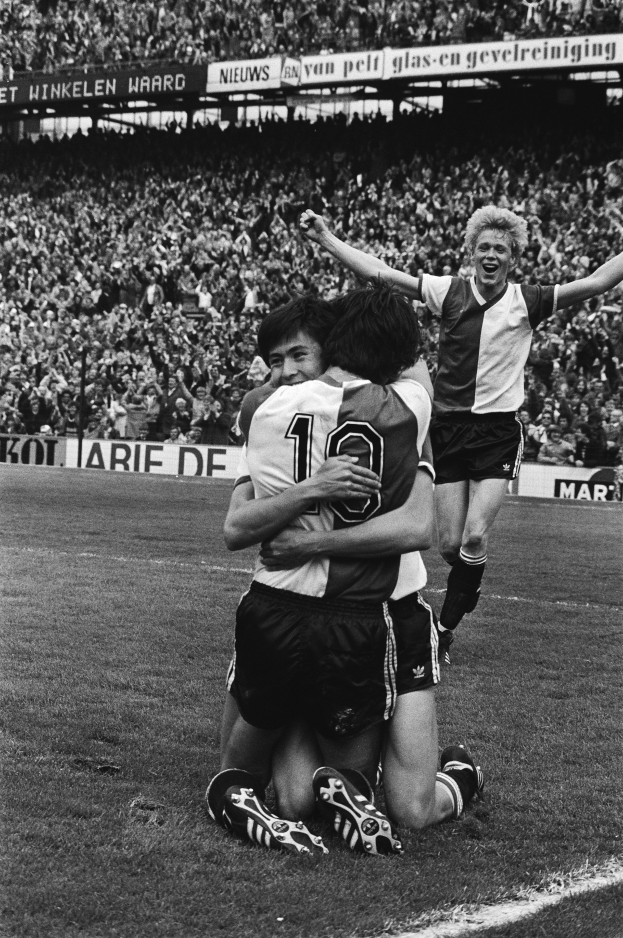 Drei Fußballspieler umarmen sich auf dem Boden, feiern ein Tor vor einer jubelnden Menge in einem schwarz-weißen Stadion.
