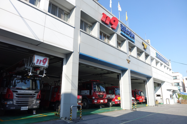 Feuerwache mit mehreren Feuerwehrautos vor dem Gebäude geparkt, Gebäude mit Fenstern, Säulen und Namensschildern, Fahnen mit Stangen im Vordergrund, andere Gebäude und klarer blauer Himmel im Hintergrund.