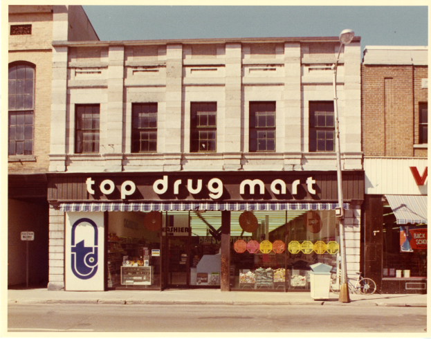 Top-Drogenmart-Ladenfront an einer Straßenecke mit einem Schild, einem Fahrrad in der Nähe, einem Mülleimer, einem Straßenschild und einem bewölkten Himmel.