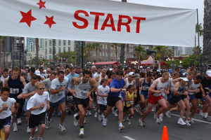 Gruppe von Läufern bei einem Marathon mit einem Verkehrskegel im Vordergrund und einer Banner mit Text im Hintergrund, umgeben von Bäumen, Laternenmasten, Gebäuden und einem klaren blauen Himmel.
