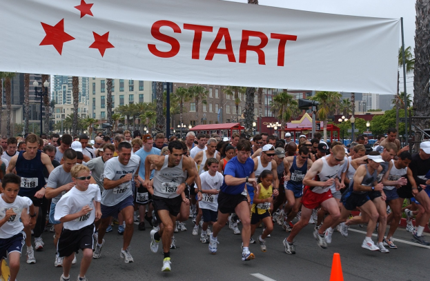 Gruppe von Läufern bei einem Marathon mit einem Verkehrskegel im Vordergrund und einer Banner mit Text im Hintergrund, umgeben von Bäumen, Laternenmasten, Gebäuden und einem klaren blauen Himmel.