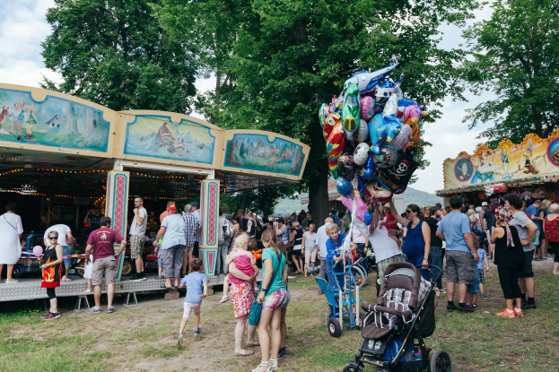 Eine Menschenmenge umgibt ein buntes, beleuchtetes Karussell auf einem Volksfest, einige halten Kinderwagen, mit Bäumen und einem bewölktem Himmel im Hintergrund.