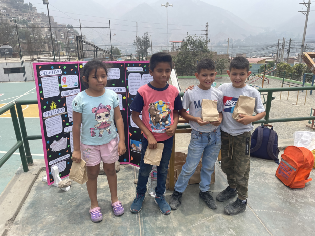 Gruppe von Kindern, die Papierbeutel auf einem Basketballfeld halten, mit Beuteln und Gegenständen auf dem Boden, einer Tafel mit Text links und Hintergrundelementen wie Zaun, Pfosten, Straßenlaternen, Strommasten, Bäumen, Gebäuden, Hügeln und Himmel.