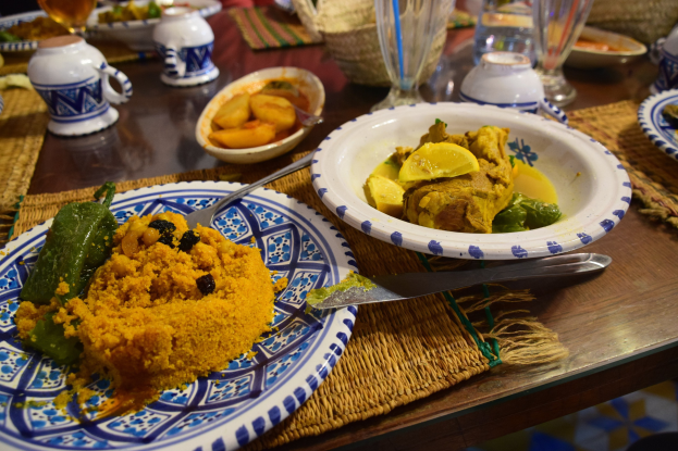 Ein Tisch mit Tellern voller Essen, Weingläsern, Schalen, Krügen, Besteck und Matten, mit Menschen im Hintergrund bei einem traditionellen marokkanischen Mahl.