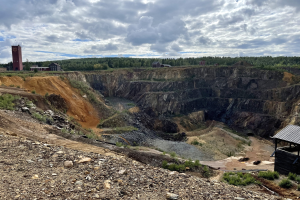 Großer Tagebau auf einem Schotterfeld mit verstreuten Steinen und Pflanzen, umgeben von Bäumen, Gebäuden und einem Schuppen unter einem bewölkten Himmel, stellvertretend für die kanadische Bergbauausweitung.