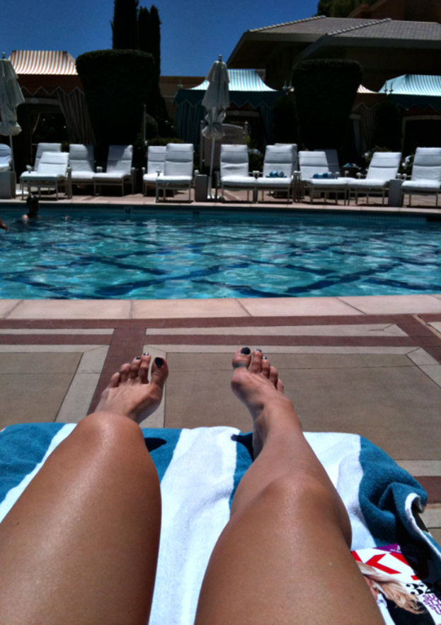 Frauen auf einem Poolbett, mit einem Swimmingpool und mehreren anderen Poolbetten mit Handtüchern im Hintergrund, sowie Büschen und Häusern.