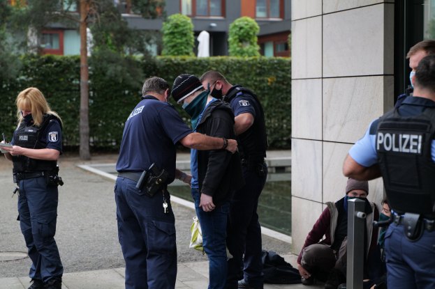 Eine Gruppe von Polizeibeamten steht vor einem Gebäude, während eine Frau ein Buch und einen Stift hält, mit Menschen in der Nähe und Grünzeug im Hintergrund.