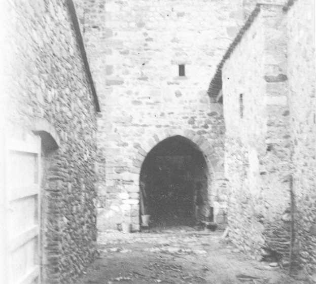 Schwarzes und weißes Foto eines alten Steingebäudes mit einem zentralen Bogengang, flankiert von Mauern, beschriftet mit "Der Eingang zur Burg" unten.