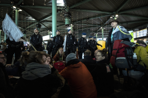 Eine Gruppe von Menschen steht vor einer protestierenden Menge in einem Bahnhof, mit Säulen und Deckenlampen im Hintergrund.