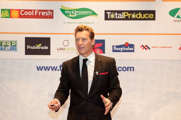 Ein Mann in einem schwarzen Blazer steht und hält ein Objekt, während er spricht, mit einem weißen Banner und einer weißen Tafel mit Organisationslogos im Hintergrund.