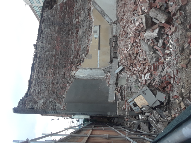 Teilweise abgerissenes Gebäude mit verstreuten Ziegeln und Metallstangen, andere Gebäude und einen klaren blauen Himmel im Hintergrund.