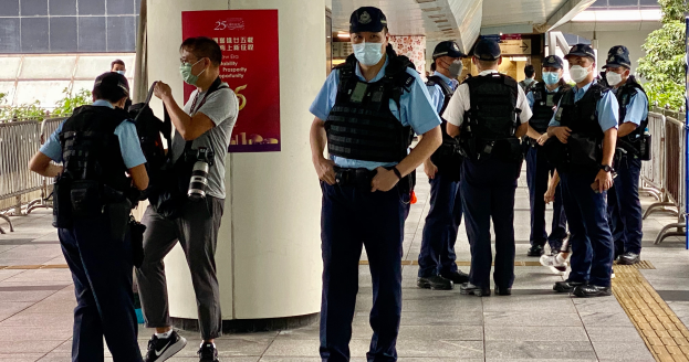 Eine Gruppe uniformierter Polizeibeamter, einige mit Kameras, steht in einer Reihe vor einem Gebäude mit einem Banner an einer Säule, Geländern, Pflanzen, Bäumen und Deckenleuchten im Hintergrund.