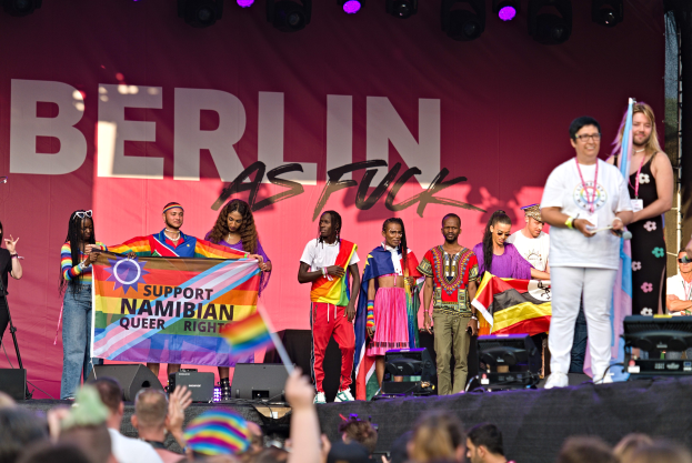 Gruppe von Menschen auf einer Bühne mit einem Banner, auf dem "Berlin als Fuck" steht, mit Lautsprechern, Mikrofonen und einer feiernden Menge unten.